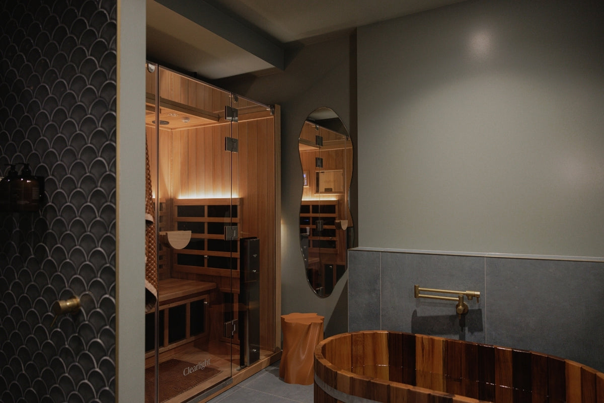 A modern bathroom interior featuring a wooden hot spring tub (onsen) in the foreground and a glass-enclosed wooden sauna in the background, with gray walls and a dark patterned tile wall.