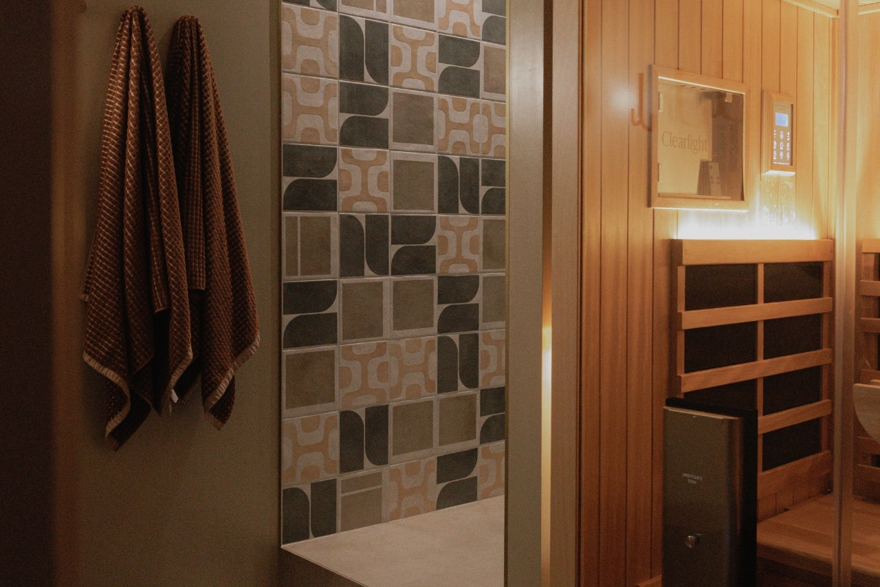 A modern sauna room interior with patterned mosaic wall tiles, hanging brown towels, wooden shelving, and warm ambient lighting.